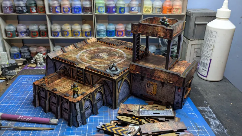 Wall-mounted wooden shelves hold rows of small paint jars. Labels on the jars read “TALI’S PAINTS,” “FLAME,” “SAND,” “COLOUR,” “CITADEL COLOUR,” “LAVA FLOW,” “PINK HORROR,” “CHROME,” “ULTRAMARINE BLUE,” “GREEN,” “PURPLE,” “ORANGE,” and “BROWN.” Below the shelves, a weathered multi-level terrain piece features arched openings, rusted metal textures, hazard-striped edges, and a circular platform bearing a logo. Two miniature figurines stand on the terrain: one in green armor on the upper platform and one in brown armor near the base. In front of the terrain, a grey plastic carrying case with a clear lid lies open on a blue-tiled surface.