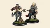Two detailed miniatures from the new Lumberjacks team for Guild Ball are displayed. On the left, a character with a beard and a red scarf stands confidently next to a small stack of logs, holding a large axe. On the right, a muscular figure with blonde hair and a serious expression wields a similar axe, dressed in dark clothing with armor accents. Both figures are mounted on circular bases with green grass detailing.