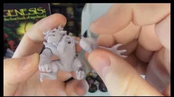 A person holds a detailed miniature of a creature from Fenris Games' Wyrdworld series, showcasing its intricate features such as muscular limbs and a fierce expression. The miniature is unpainted and made of a light-colored material, emphasizing its sculpted details. In the background, a colorful game box titled "GENESIS: The First Book of Nightmares" is partially visible, along with other miniatures displayed on a shelf.