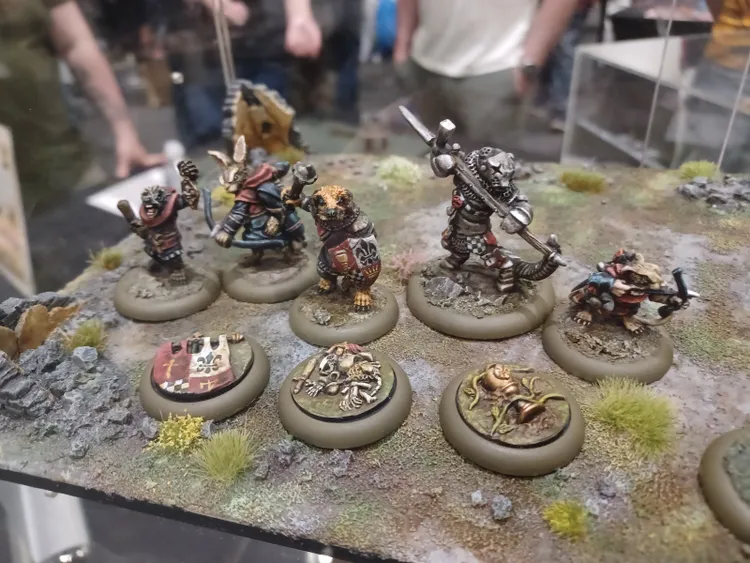 A group of anthropomorphic animal miniatures stands on a textured, grassy base, showcasing a variety of characters from the Burrows and Badgers range. The figures include a rabbit wielding a staff, a bear in armor holding a weapon, and several other creatures, each adorned with unique details and vibrant colors. Surrounding them are circular bases featuring thematic elements like skeletons and plants, enhancing the fantasy setting.