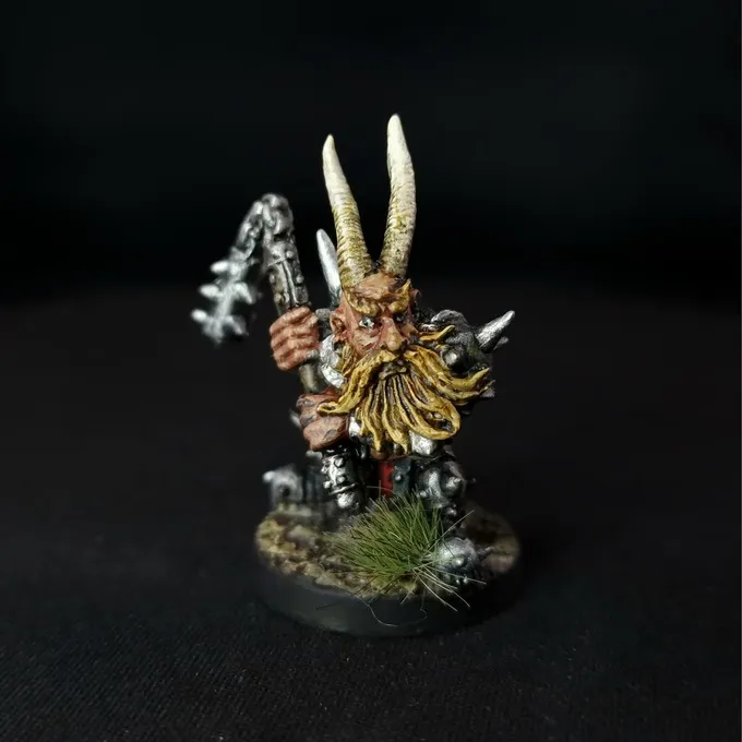 A painted miniature of a Chaos Dwarf with a long yellow beard, wearing metallic armor and a horned helmet. The figure holds a staff with a twisted design and stands on a circular base with small patches of grass and rocks. The background is black.