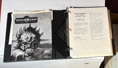 A three-ring binder containing the playtest manuscript for "ManaStorm: The Magic Adventure Game," featuring a black and white cover illustration of a character with a spiked helmet and a weapon. The title "MANASTORM The Magic Adventure Game" is prominently displayed at the top, along with the version number and date on the first page inside the binder. Additional pages include loose character sheets and player handouts, indicating the game's mechanics and spells.