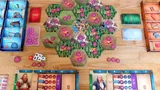 Board game setup with hexagonal green terrain tiles featuring pink and purple structures. Miniature figures and small tokens scattered across. Two player boards with character portraits, resource dials, and action icons. Dice and small bags of colored tokens on the table.