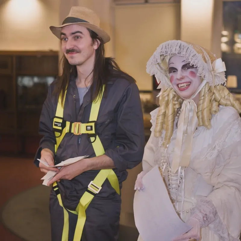 A man dressed in a dark jumpsuit with bright yellow safety straps and a beige hat stands next to a woman in an elaborate white Victorian-style dress with lace and ruffles, her hair styled in long, curled locks. Both are smiling and holding papers, with the backdrop featuring the interior of a gothic building, likely part of the event's vampire-themed atmosphere.