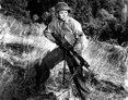 A black-and-white image features a soldier in a military uniform, wearing a helmet and a sergeant's insignia, crouched in a grassy field. He holds a machine gun with both hands, poised as if ready for action. The background consists of blurred greenery, suggesting a natural setting.