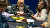 A man with glasses and a beard wearing a black Pac-Man-themed shirt plays a board game with colorful tokens on a blue and yellow game board. A child reaches toward the game. The game box has text including "STEM" and "Space".