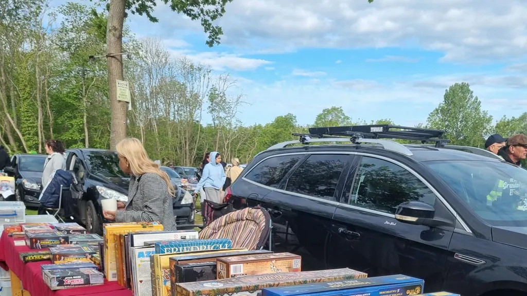 Outdoor car boot sale with red tablecloths displaying board games, DVDs, and books. People browsing and parked cars in background.