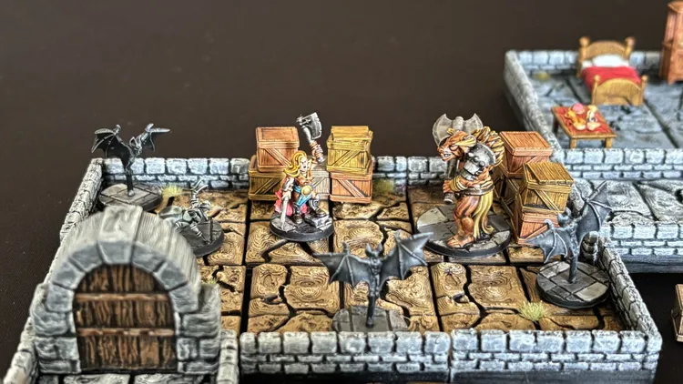 A detailed tabletop scene features a modular dungeon made up of textured floor tiles, with stone walls and wooden crates scattered throughout. In the center, a heroic character with long blonde hair and a battle axe stands ready, facing a menacing creature resembling a minotaur. Nearby, a bat figure hovers, adding to the atmosphere of adventure and danger in this 15mm scale dungeon-crawl setting.