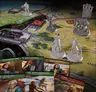 A detailed board game scene features a colorful map depicting the land of the Two Rivers, with various regions and locations marked. Prominent on the map are several unpainted miniatures representing characters from The Wheel of Time, including a figure holding a spear and another with a staff, alongside a horse. In the foreground, several character cards are displayed, showcasing allies such as Sheriam Bayanar and Bain and Chiad, each with distinct artwork and names.