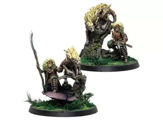 A pair of intricately detailed miniatures from Corvus Belli, featuring a character with long, wild hair wielding a spear, accompanied by a large, stylized creature resembling a lion with a mane of flames. The figures are set on a base adorned with lush green grass and a gnarled tree, enhancing the naturalistic theme. The character appears to be in a dynamic pose, suggesting action or readiness, while the creature stands alert beside them.