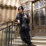 A person dressed in a black leather jacket and dark clothing stands confidently on a staircase, striking a rock-and-roll pose with one hand raised in a gesture. They wear sunglasses and have a stylish, short hairstyle, complemented by accessories like chains and rings. In the background, ornate wooden doors with decorative ironwork are visible, hinting at the gothic architecture of the venue.