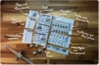 Six printed game sheets on wooden surface. Labels: "first combat," "My gold poise," "Scribe and their scrolls," "Apothecary and their potions," "Marie-Agnes and her equipment," "Final Boss." Black-and-white illustrations of characters, items, and dice mechanics. Five dice on left.