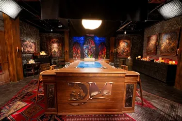 A richly designed tabletop setting features a large, intricately carved wooden table at the center, adorned with a blue surface that suggests a gaming area. Surrounding the table are various chairs, and the backdrop includes stone walls with colorful stained glass windows, casting vibrant light across the scene. The space is decorated with candles, artwork depicting fantasy themes, and a patterned rug, creating an immersive atmosphere suitable for a Dungeons & Dragons actual play show.