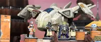 A detailed scene features a futuristic spaceship with a sleek design, prominently displayed in the background. In the foreground, several 28mm scale miniatures are arranged, including a female pilot in an orange jumpsuit, two armed security officers in dark uniforms, a large alien figure adorned in ornate armor, and a small robot. The setting includes wooden crates, enhancing the sci-fi atmosphere.