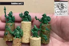A collection of five green goblin miniatures is displayed on cork bases, showcasing their unique designs and poses. The goblins are equipped with various weapons, including spears and axes, and wear distinct armor pieces, highlighting their mischievous character. The background is neutral, allowing the vibrant green of the miniatures to stand out prominently.