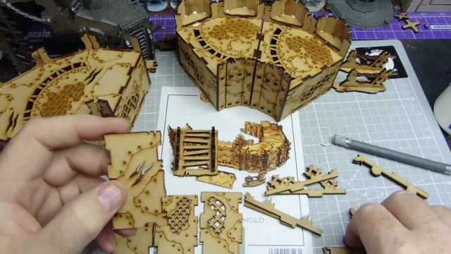 A hand holds a small, intricately cut piece of laser-cut wood, featuring detailed patterns and textures. Surrounding the hand are larger assembled sections of similar wooden terrain pieces, showcasing various designs, including grids and panels. In the background, a printed layout with additional terrain components is visible, indicating a crafting or gaming setup.