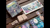 A wooden table displays the board game Nakasendo. The game features a modular map with floral and mountain-themed tiles, wooden pieces, and numbered cards. Visible text on cards includes "24," "25," and "26." Small round tokens and a dice are scattered nearby.