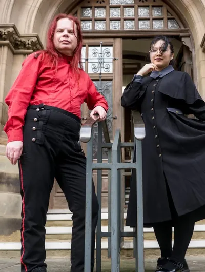 Two individuals pose in front of a grand, gothic-style building. One person, wearing a bright red shirt and black trousers with red stripes, stands confidently with arms crossed. The other person, dressed in a long black coat with a high collar and a blue shirt underneath, holds the hem of their coat, creating a dramatic silhouette. The ornate entrance of the building, featuring large wooden doors and intricate stonework, serves as a striking backdrop.