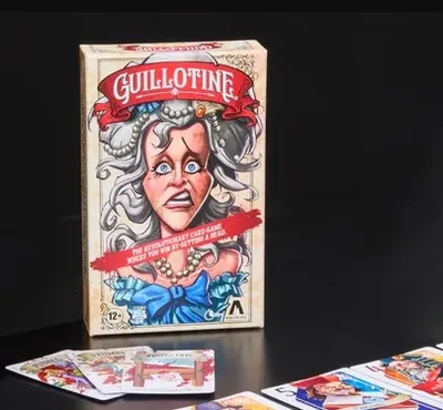 The image features the box of the card game "Guillotine," prominently displaying an illustration of a startled woman with gray hair and a blue bow, set against a light-colored background. Next to the box, a wooden guillotine prop is shown, designed to hold the noble cards. In the foreground, several tarot-sized noble cards are laid out, showcasing colorful illustrations of various characters, each with distinct expressions and attire, reflecting the game's theme of the French Revolution.