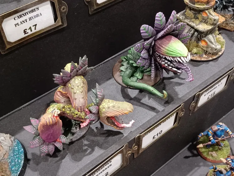 A display of intricately painted fantasy miniatures features several man-eating plants, showcasing vibrant colors and detailed designs. The plants have multiple heads, with some exhibiting bright greens and purples, while others display a mix of pinks and yellows. The miniatures are arranged on a dark surface, with price tags visible next to them, highlighting their unique and imaginative designs.