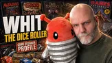A man with a shaved head and a beard is holding a plush toy resembling a red creature with large eyes and a segmented silver body. He is wearing a dark green sweater and has a serious expression. In the background, there are various tabletop gaming items, including a magazine titled "Games Unplugged" and a box for "Dark Conspiracy 2nd Edition." The overall scene conveys a blend of gaming culture and personal connection to the hobby.