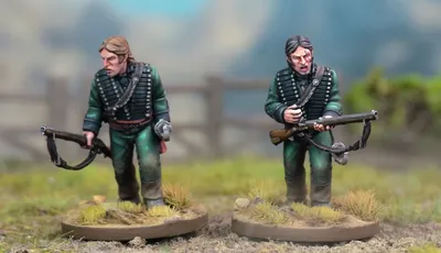 Two detailed miniatures of soldiers from the Sharpe’s Rifles series are displayed on textured bases, set against a blurred natural background. Both figures wear green uniforms with black crossbelts; one holds a rifle and has a serious expression, while the other appears to be in a more contemplative pose. The bases feature tufts of grass, enhancing the realistic terrain effect.