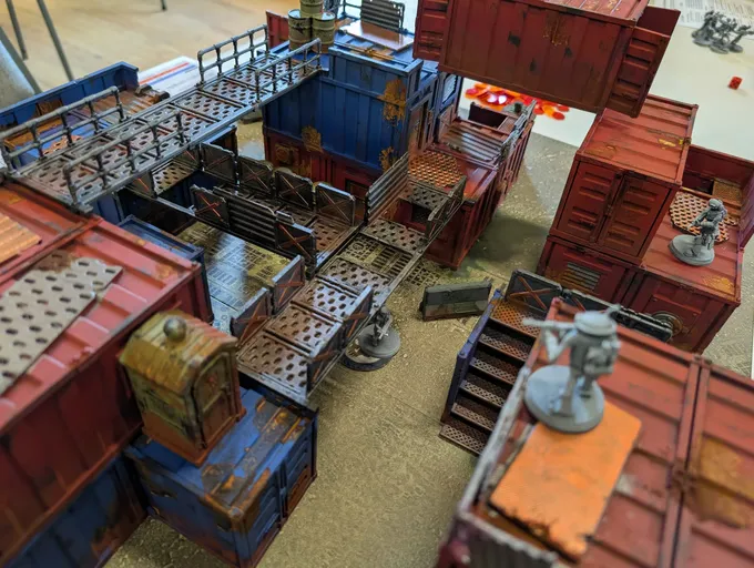 Detailed miniature model of a multi-level industrial or sci-fi terrain setup. The scene features interconnected shipping containers and modular structures stacked and arranged in a compact layout. The containers and buildings are painted in various colors, including blue, red, brown, and gray, with some showing weathering effects.

A network of metal walkways and staircases connects the different levels, with railings along the edges. Some walkways have gridded or perforated flooring. Several small details are visible, including a white water cooler or dispenser with a cup on a platform, a small stool, and scattered debris or equipment on the ground. A ladder is attached to one of the containers. No text or signage is legible in the image.