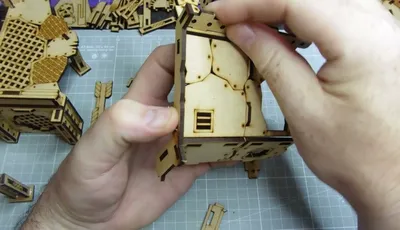 A hand holds a piece of laser-cut wood, part of the TT Combat Orc Mega Bunker model, showcasing intricate details like a small window and textured surfaces. Surrounding the hand are various other wooden components and parts of the model, laid out on a cutting mat with grid lines. The overall scene emphasizes the assembly process of the Orc Sektor level 2 build.