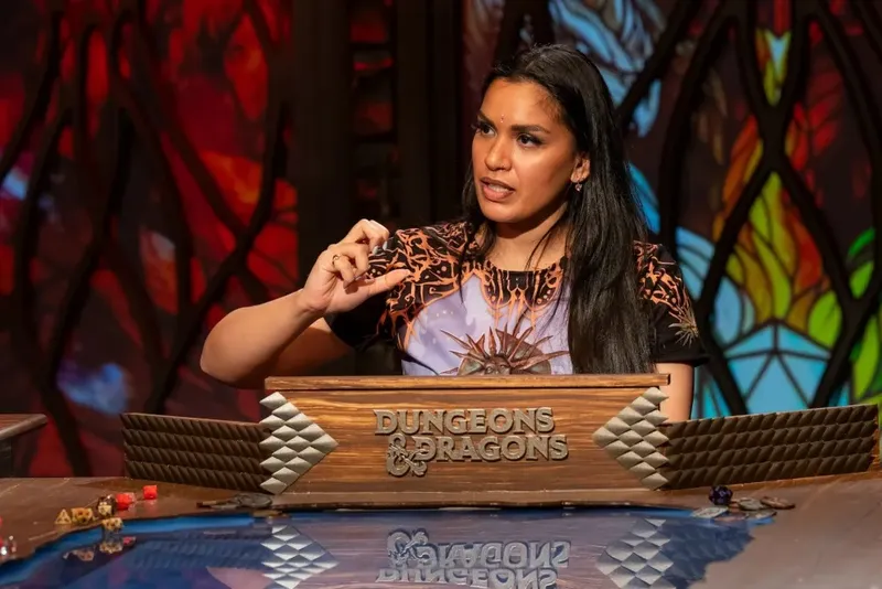 A woman with long black hair is animatedly speaking while gesturing with her right hand, seated at a table adorned with a "Dungeons & Dragons" sign. She wears a colorful shirt featuring a sun design and is positioned in front of a backdrop of stained glass, which adds a vibrant and atmospheric touch to the scene. The table is decorated with various gaming accessories, enhancing the tabletop roleplaying game setting.