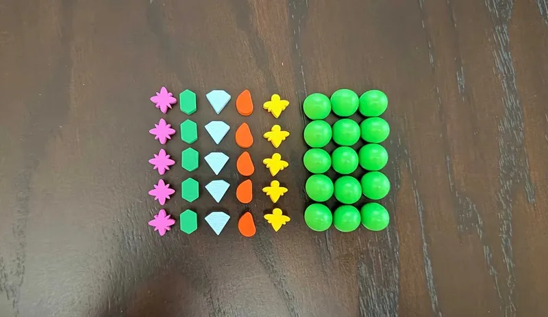 A collection of game components for the Wingspan: Americas Expansion is displayed on a dark wooden surface. The pieces include various shapes and colors: pink flowers, green hexagons, blue diamonds, orange teardrops, yellow birds, and multiple green spheres arranged in a grid. The vibrant colors and distinct shapes suggest they are used for gameplay mechanics related to the new expansion.