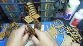 A workspace filled with various pieces of laser-cut terrain and building components, primarily made of light brown wood. In the foreground, a hand is assembling parts, including a set of stairs and wall sections, while a blue cutting mat with grid lines provides a base. In the background, several painted structures are stacked, alongside bottles of glue and other crafting supplies.