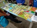 A tabletop game setup is displayed on a light blue table, featuring a variety of colorful game pieces and cards. The game board is filled with hexagonal tiles, tokens, and cards in shades of green, yellow, red, and brown, indicating different resources and actions. In the foreground, a person wearing an orange sweater and black pants is seated, with a blue backpack visible beside them, while another player’s hands are seen above the game board.