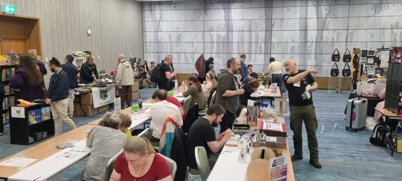 A bustling convention scene at Gaelcon features a variety of attendees engaged in different activities. In the foreground, several people are seated at tables, painting miniatures and playing games, while others browse merchandise at vendor booths in the background. The room is well-lit, with a modern design and a large mural depicting a forest scene on the wall.