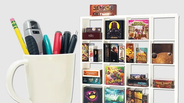 A white ceramic cup filled with pens and a stapler sits on the left. To the right is a white grid-style shelf holding multiple small board game boxes, including visible titles such as "GLOOMHAVEN", "MARS", "POTION EXPLOSION", "DICED VEGGIES", "TERRAFORMING MARS", "CUBITOY", "Sleeping Gods", and others.