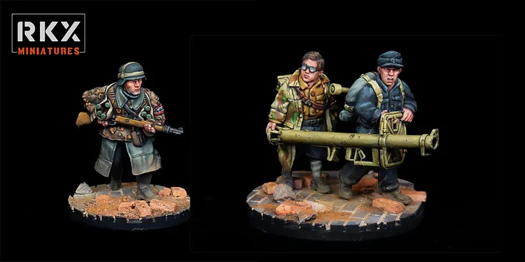 A detailed miniature scene features two German soldiers, one operating a Panzerschreck anti-tank weapon, while the other assists in carrying it. The soldier on the left wears a uniform with a distinctive insignia, while the one on the right is dressed in a long coat and goggles, suggesting a tactical role. They stand on a textured base with scattered bricks and debris, evoking a war-torn environment.