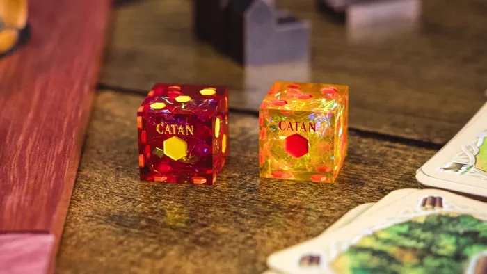 Two translucent dice with "CATAN" text and colored internal shapes on a dark surface.