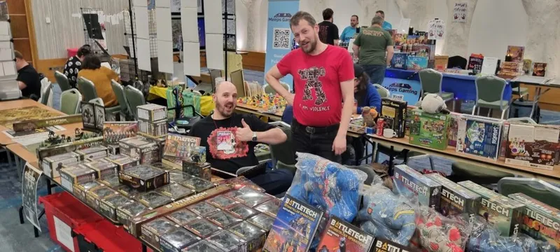 Two men are standing behind a table filled with various tabletop gaming products, including multiple boxes of "BattleTech" miniatures and other games. One man, wearing a red shirt with a graphic design, is giving a thumbs-up, while the other, in a black shirt, smiles at the camera. In the background, other attendees are engaged in gaming activities, with colorful game boxes and figures visible on nearby tables.
