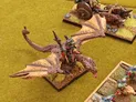 A detailed miniature scene features a dragon with outstretched wings, carrying a rider adorned in colorful armor and wielding a spear. In the background, a cart pulled by two boar-like creatures is visible, along with a grassy terrain dotted with small patches of foliage. The overall setting evokes a classic Oldhammer aesthetic, showcasing the vibrant colors and intricate details typical of tabletop gaming miniatures.