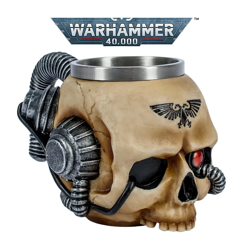 Beige ceramic mug shaped like a stylized human skull with mechanical augmentations. A metal rim lines the top of the mug. Two large, cylindrical, metallic mechanical components resembling pistons or servos are attached to the sides of the skull, connected by coiled black cables. A black eagle emblem with outstretched wings and a skull in the center is tattooed or printed on the forehead. The background shows a metallic banner with the text "WARHAMMER 40,000" in bold, futuristic lettering. The banner has a red and white stripe along the bottom edge.