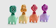 Three ceramic elephant-shaped containers with metallic tops. Left: cream elephant, gold top with abstract swirl design. Center: peach elephant, red top with abstract geometric design. Right: lavender elephant, purple top with abstract circular design. All on white background.