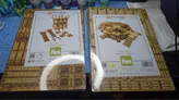 Two boxes of TT Combat tabletop scenery are displayed on a work surface. The left box is labeled "Sector 4 Storage Tanks," featuring images of storage tanks, ladders, and crates. The right box is labeled "Sector 4 Mega Turbine," showcasing a detailed turbine design. In the background, various paint bottles are visible, indicating a workspace for model building and painting.