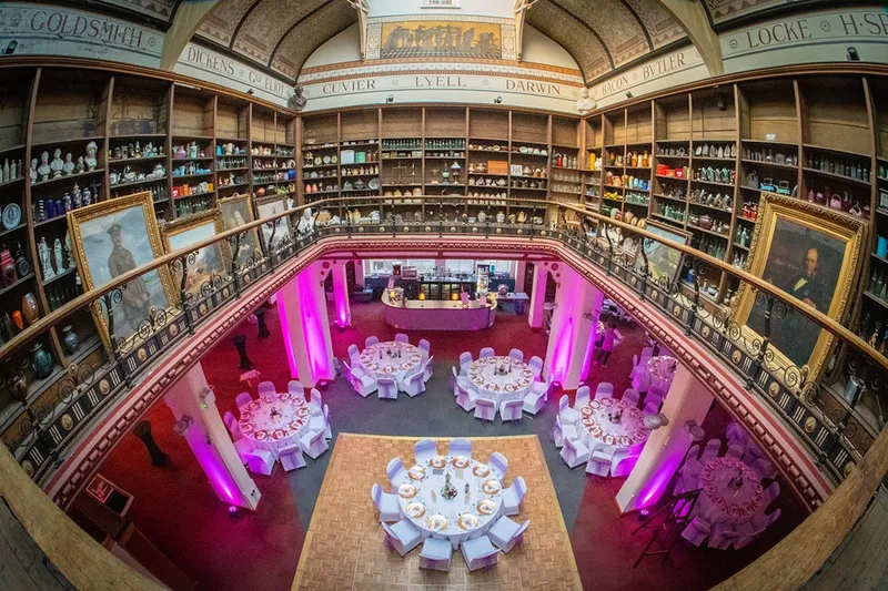 A spacious, elegantly decorated venue features several round tables set for dining, each adorned with white tablecloths and plates. The room is illuminated with soft pink lighting, highlighting the ornate architecture, including a high ceiling and shelves filled with various decorative items. In the background, a bar area is visible, and a large portrait hangs on the wall, adding to the gothic atmosphere suitable for the Vampire Games Day II event.