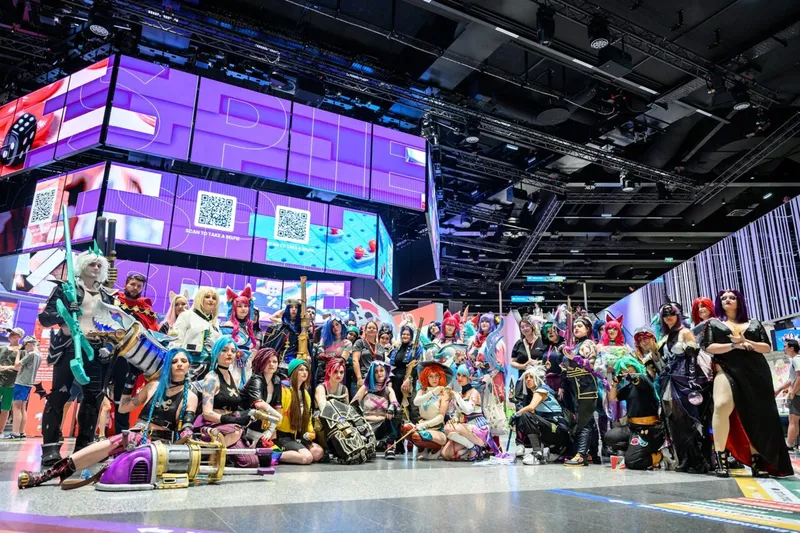 A large group of cosplayers is gathered in a spacious venue, showcasing a variety of colorful and intricate costumes inspired by different characters, primarily from video games. Behind them, large digital screens display vibrant graphics and event information, contributing to the lively atmosphere. The setting is well-lit, with a modern design that enhances the excitement of the gaming event.