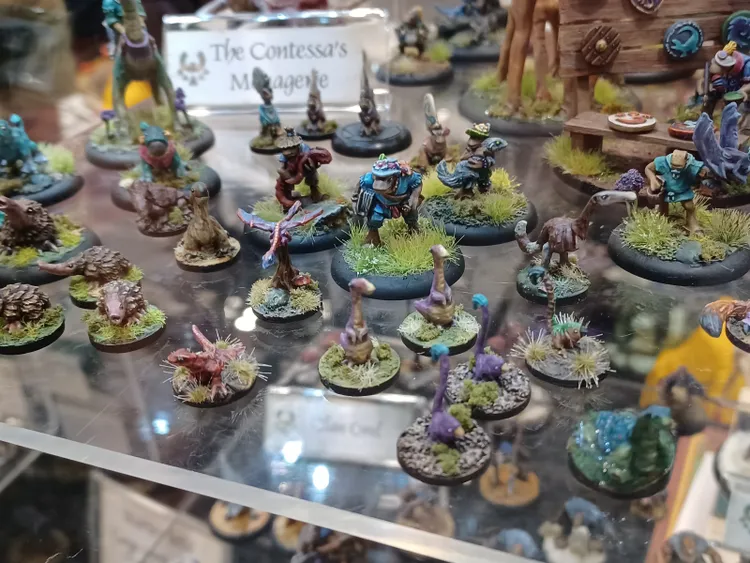 A diverse array of fantasy miniatures is displayed, featuring various anthropomorphic characters and creatures. In the foreground, small figures include a frog-like warrior in blue armor, several colorful, small dinosaurs, and whimsical man-eating plants, all set on bases with tufts of grass. The background showcases larger figures, including a tall creature and a sign reading 
