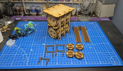 A detailed assembly of the TT Combat Sector 4 Storage tanks is displayed on a blue cutting mat marked with a grid. The main structure consists of two large, cylindrical tanks stacked vertically, supported by a wooden frame with intricate detailing. Surrounding the tanks are various components, including two sets of ladders, several hazard markers labeled "DANGER," and additional structural pieces, all crafted from a light wood material. In the background, a miniature figure and a globe stand are visible, adding context to the tabletop gaming environment.