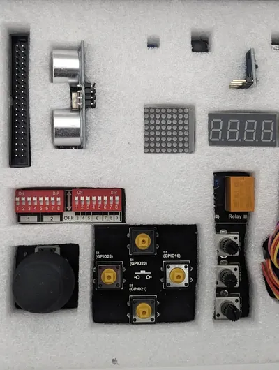 A collection of electronic components is neatly arranged on a white foam background. Key items include a pair of ultrasonic sensors, a digital display, various buttons, a relay, and a DIP switch. Additional components like a rotary encoder and several connectors are also present, showcasing a variety of parts typically used in DIY electronics projects.