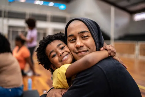 A joyful scene captures a smiling child with curly hair embracing an adult, who is also smiling and wearing a hoodie. They are in a spacious indoor setting, likely a gym or community center, with other people in the background engaged in various activities. The warm expressions and close interaction convey a sense of love and connection between them.
