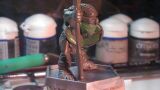 A small, detailed miniature figure stands on a square base, holding a staff and wearing a green mask. The figure is dressed in textured armor, showcasing intricate details like scales and straps. In the background, various paint pots and tools are visible, indicating a workspace for painting or modeling miniatures.