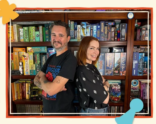 Felicia Day and Wil Wheaton standing with arms crossed in front of a bookshelf filled with board games.