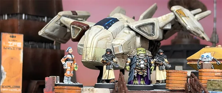 A detailed scene features a futuristic spaceship with a sleek design, prominently displayed in the background. In the foreground, several 28mm scale miniatures are arranged, including a female pilot in an orange jumpsuit, two armed security officers in dark uniforms, a large alien figure adorned in ornate armor, and a small robot. The setting includes wooden crates, enhancing the sci-fi atmosphere.